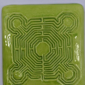 May include: A square, ceramic coaster with a maze design. The coaster is a bright, glossy green color, with a raised maze pattern in a slightly darker shade of green. The maze is centered on the coaster and has a complex, geometric design.