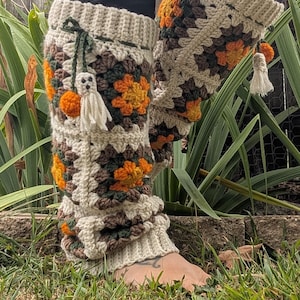 May include: Crocheted leg warmers in a granny square pattern, featuring cream, brown, orange, and green squares. Each leg warmer has a cream-colored cuff and is adorned with a small ghost and orange pom-poms. The leg warmers are designed for comfort and style.