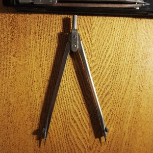 May include: A silver compass with a metal point on each end. The compass is open and ready to draw a circle.