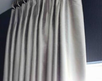 May include: A set of floor-length, light gray curtains with a subtle sheen, featuring deep vertical pleats. The fabric is suspended from a dark metal curtain rod by a series of matching circular metal rings, creating uniform folds along the length.