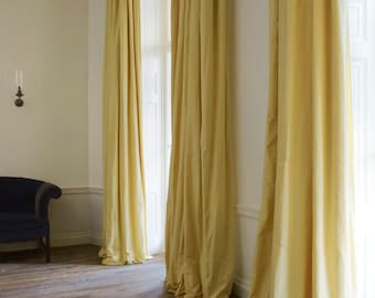 May include: Three floor-length, solid pale yellow curtains hang from a rod, extending from the ceiling to the floor. The fabric is opaque and drapes in vertical folds, covering the windows and reaching the wooden floorboards in the room.