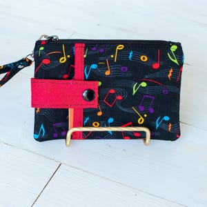 May include: A black zippered wallet with a wrist strap, featuring a colorful musical note pattern. The wallet has a red flap with a black button closure and a gold-colored metal handle.