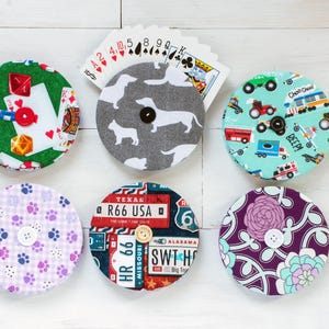 May include: Six round fabric coasters with different designs. The coasters feature a variety of patterns, including paw prints, a dachshund, a road sign, a floral design, and a train. The coasters are all made of fabric and have a button in the center.
