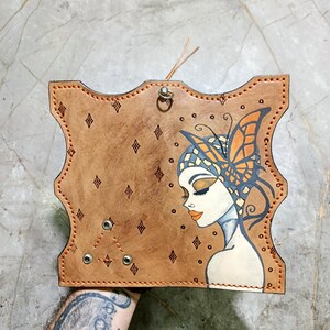 Könnte beinhalten: Handgenähte Lederbrieftasche mit einer dekorativen Illustration zum Thema Schmetterling. Die Brieftasche hat eine hellbraune Farbe mit orangefarbenen Nähten. Die Illustration zeigt eine Frau mit Schmetterlingskopfschmuck und -flügeln.