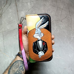 Könnte beinhalten: Eine handbemalte Ledergeldbörse mit Bugs Bunny im Smoking. Die Geldbörse hat eine Handschlaufe mit roten und rosa Akzenten. Das Design umfasst einen schwarzen Rand und einen braunen kreisförmigen Hintergrund.