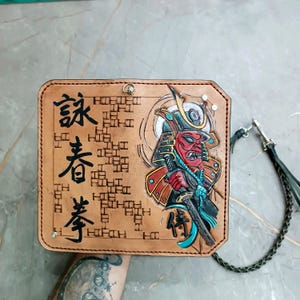 Puede incluir: Cartera de cuero color canela con un diseño en relieve de un guerrero samurái de rostro rojo con una espada y un casco. La cartera presenta caracteres japoneses y una correa de cuero trenzado con un cierre metálico.