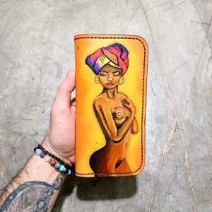 Pode incluir: Carteira de couro pintada à mão com uma mulher com um turbante colorido. A obra de arte está em um fundo amarelo. A carteira é segurada em uma mão, mostrando os detalhes intrincados e o artesanato do design.