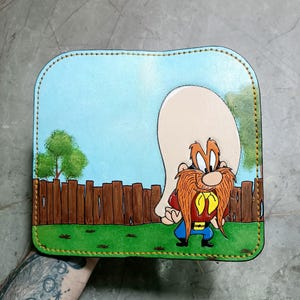 Könnte beinhalten: Eine handgefertigte Ledergeldbörse mit einer Cartoonfigur mit großem weißem Hut, rotem Schnurrbart und gelbem Hemd. Der Hintergrund zeigt einen blauen Himmel, grünes Gras und einen Holzzaun.
