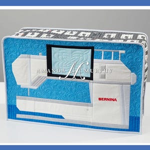May include: A custom sewing machine cover with a quilted design. The cover features a blue background with a sewing machine illustration, gray and white accents, and the text "B 990 Cover-Up". The brand name "BERNINA" is visible.