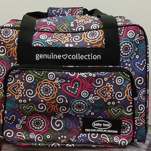 May include: A black sewing machine bag with a colorful heart and floral pattern. The bag has a black handle and a zipper closure. The bag has a label that says "genuine ♥ collection" and "baby lock for the love of sewing".