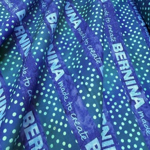 May include: Close-up of a blue fabric with white polka dots and diagonal stripes. The word "BERNINA" is printed in white on the stripes, along with the phrase "made to create". The fabric has a textured appearance.