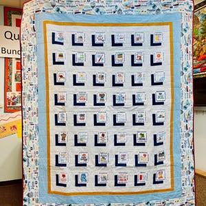 May include: A white quilt with a blue and yellow border. The quilt is made up of squares with different sayings and designs. The quilt is hanging on a wall.