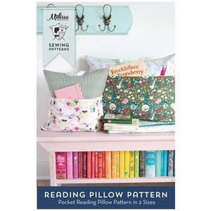May include: A sewing pattern for a reading pillow with a pocket. The pattern is called "Reading Pillow Pattern" and is available in two sizes. The image shows a pink bench with a stack of books and two pillows with pockets. One pillow has a green and white striped fabric and the other has a floral print. The text "Freckleface Strawberry" is visible on the floral pillow.