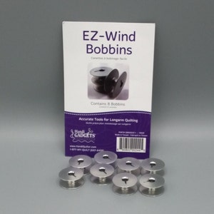 May include: A package of EZ-Wind Bobbins with the text "EZ-Wind Bobbins" and "Contains 8 Bobbins". Eight silver metal bobbins are in front of the package. The package also has the text "Accurate Tools for Longarm Quilting".