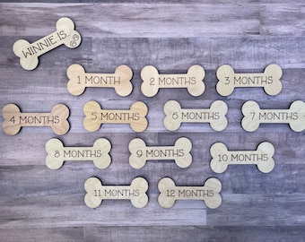 May include: Wooden dog bone-shaped milestone markers. One bone reads "WINNIE IS..." with paw prints. The other bones are labeled "1 MONTH" through "12 MONTHS". These are perfect for tracking a puppy's growth.