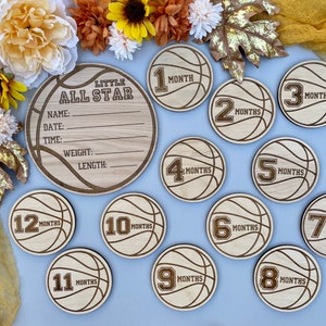 以下が含まれることがあります： バスケットボールをテーマにした木製ベビーマイルストーンセット。セットには、「Little All Star」と書かれた大きな丸いプレートと、赤ちゃんの名前、日付、時間、体重、身長を記入するスペースがあります。バスケットボールの形をした小さな丸いプレートが12枚あり、それぞれに1から12までの数字と「月」の文字が書かれています。