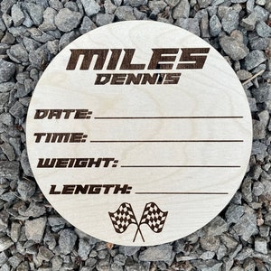 K&ouml;nnte beinhalten: Eine runde Holzplakette mit dem Namen "MILES DENNIS" in schwarzer Schrift. Die Plakette hat Platz zum Eintragen von Datum, Uhrzeit, Gewicht und L&auml;nge. Sie zeigt eine Grafik mit einer karierten Flagge.
