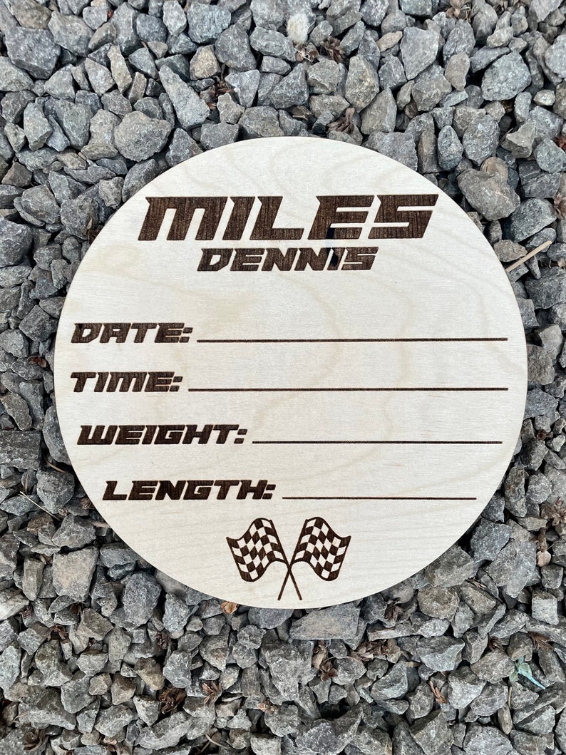 K&ouml;nnte beinhalten: Eine runde Holzplakette mit dem Namen "MILES DENNIS" in schwarzer Schrift. Die Plakette hat Platz zum Eintragen von Datum, Uhrzeit, Gewicht und L&auml;nge. Sie zeigt eine Grafik mit einer karierten Flagge.