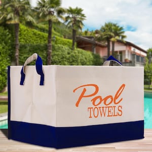 Könnte beinhalten: Eine rechteckige Canvas-Tragetasche mit den Worten "Pool TOWELS" in orangefarbener Schrift. Die Tasche hat einen weißen Korpus, einen marineblauen Boden und marineblaue Griffe mit weißem Futter. Im Hintergrund sind ein Pool und Palmen zu sehen.