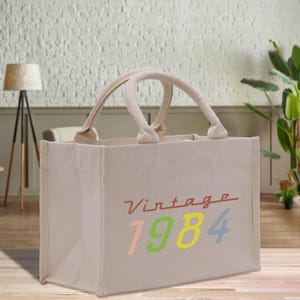 May include: Beige tote bag with two handles. The bag features the text "Vintage 1984" in a colorful design. The word "Vintage" is in red, and the numbers are in pink, green, yellow, and blue.