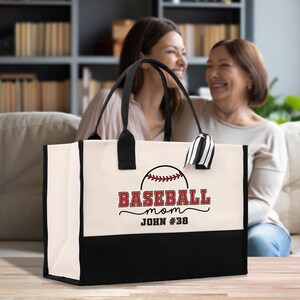 May include: A canvas tote bag with a black bottom and handles. The bag features a baseball graphic with the words "BASEBALL mom" in red and black, along with a name and number. The bag is cream-colored with black accents.