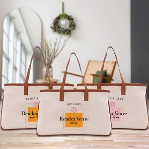 Könnte beinhalten: Drei Canvas-Tragetaschen mit braunen Ledergriffen und -besatz. Jede Tasche hat ein Champagnerflaschen-Etikettendesign mit dem Text "Rendez Veuve" und anderem Text. Die Taschen sind cremefarben mit hellbraunem Besatz.
