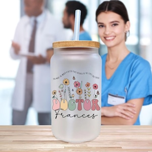 May include: Frosted glass tumbler with a bamboo lid and a clear straw. The tumbler features a floral design with the word "DOCTOR" in colorful letters and the name "Frances" below. The text "YOU MAKE THE WORLD A BETTER PLACE BECAUSE OF THE LIVES YOU TOUCH" is printed on the tumbler.