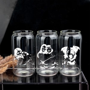 Peut inclure: Trois verres transparents en forme de canette avec des motifs blancs gravés personnalisés. L'un représente un couple, un autre un autre couple et le troisième un chien. Les verres sont présentés sur une surface transparente sur fond noir.
