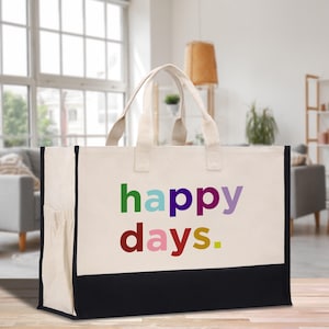 May include: A large tote bag with a cream-colored body, black bottom, and cream-colored handles. The words "happy days." are printed in colorful letters on the front. The bag has a pocket on the side and is sitting on a wooden surface.