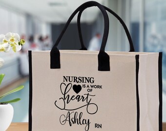 Puede incluir: Un bolso tote de lona beige con ribetes y asas negras. El bolso presenta el texto "NURSING IS A WORK OF HEART Ashley RN" en escritura negra y un gráfico de corazón. El bolso está diseñado para transportar objetos.
