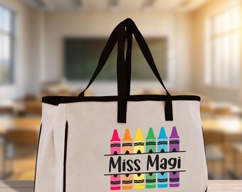 May include: A beige tote bag with black trim and handles. The front features a row of colorful crayon illustrations above the text "Miss Magi" in a black script font. The bag is designed for carrying items.
