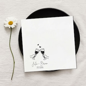 May include: A white card with a black illustration of two wine glasses clinking, with two hearts above. The card reads "Julie + Brian" and "11.11.2026" in script. The card is on a black plate, with a daisy.