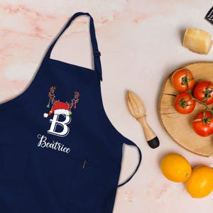 Puede incluir: Delantal azul marino con un diseño navideño. Presenta un gorro de Papá Noel rojo con cuernos y la letra "B" en blanco, junto con el nombre "Beatrice". El delantal tiene un bolsillo y una correa ajustable para el cuello.