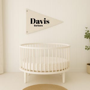 May include: A cream-colored baby crib with a round design and vertical bars. Above the crib is a cream-colored pennant banner with the name "Davis" and the name "Barbara" printed in black. The crib is on a light-colored wooden floor.