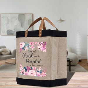 May include: A beige tote bag with black trim and leather handles. The bag features a floral design with pink and purple flowers and the text "Come unto Christ and be Perfected in Him." The bag is made of a durable material.