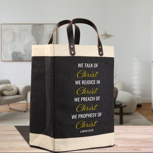 May include: A black tote bag with cream-colored trim and handles. The bag features the text "WE TALK OF Christ, WE REJOICE IN Christ, WE PREACH OF Christ, WE PROPHESY OF Christ" in white and gold lettering. The scripture reference 2 NEPHI 25:26 is also included.