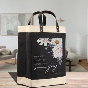 May include: A black tote bag with cream trim and leather handles. The bag has a floral design with white and pink flowers and the text "MEN ARE THAT THEY MIGHT HAVE joy".