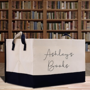 May include: A rectangular storage tote with a cream-colored body and a navy blue base. The tote has navy blue handles and the words "Ashley's Books" are written in a cursive font on the front. The background is a library.