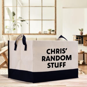 May include: A large, rectangular storage bin with a cream-colored body and a navy blue base. The bin has navy blue handles and the words "CHRIS' RANDOM STUFF" printed in bold black letters on the front.