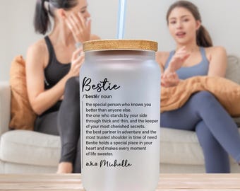 Op de afbeelding: Matglazen tumbler met een bamboe deksel en een herbruikbaar rietje. De tumbler heeft het woord "Bestie" en een definitie, samen met de naam "Michelle". De achtergrond toont twee mensen.