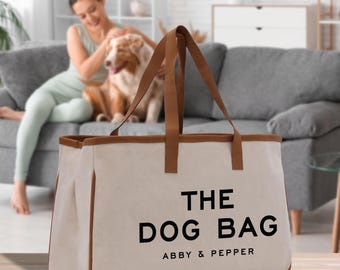 May include: A canvas tote bag with brown trim and handles. The bag is cream-colored with the words "THE DOG BAG" printed in black, along with "ABBY & PEPPER" underneath. The bag is rectangular and appears to be sturdy.
