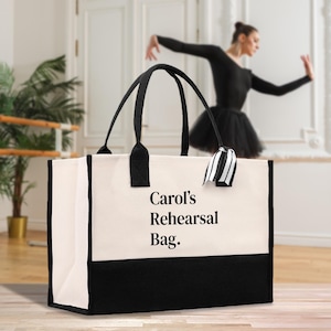 Könnte beinhalten: Eine große Tragetasche mit schwarzen Henkeln und einem schwarzen unteren Panel. Die Tasche ist cremefarben und trägt den schwarzen Aufdruck "Carol's Rehearsal Bag". Ein schwarz-weiß gestreiftes Band ist an einem Henkel befestigt.