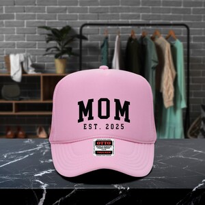 May include: A pink trucker hat with the word "MOM" in bold black letters, with "EST. 2025" below. The hat has a mesh back and a curved brim. A small "OTTO" label is on the front.