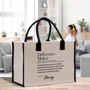Może przedstawiać: Beżowa torba typu tote z czarnymi wykończeniami i uchwytami. Torba zawiera tekst "Difference Maker" z definicją i imieniem "Nancy". Torba jest przeznaczona do przenoszenia przedmiotów.
