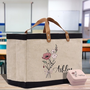 May include: A beige tote bag with a black trim and brown leather handles. The bag features a floral design with pink and white flowers and the name "Ashley" in script. A matching small pink box is next to the bag.