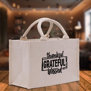 May include: A beige canvas tote bag with two handles. The bag features the words "thankful, GRATEFUL, blessed" in black script. The bag is sitting on a wooden surface.