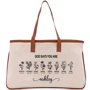 A beige canvas tote bag with a brown leather strap and the text "GOD SAYS YOU ARE" printed on it. Below the text, there are illustrations of flowers with descriptions of the corresponding Bible verse titles.