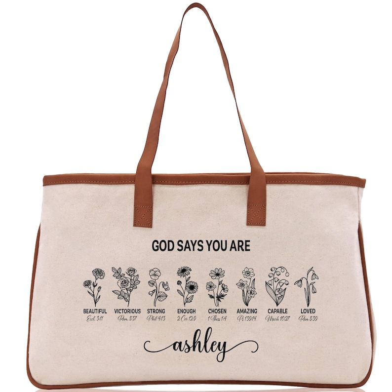 A beige canvas tote bag with a brown leather strap and the text "GOD SAYS YOU ARE" printed on it. Below the text, there are illustrations of flowers with descriptions of the corresponding Bible verse titles.