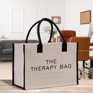 May include: A cream-colored tote bag with black trim and handles. The text "THE THERAPY BAG" is printed on the front, along with "SPEECH PATHOLOGY" and a name. The bag is rectangular and appears to be made of canvas.
