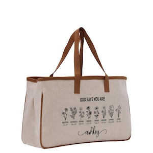 A beige canvas tote bag with a printed design of flowers and the text "God says you are Ashley" printed on it.